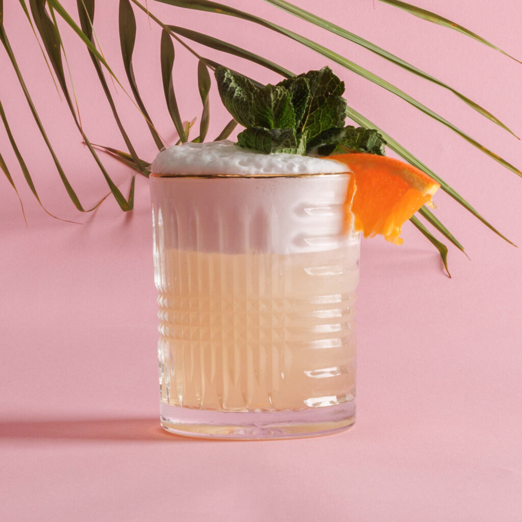 Mai Tai cocktail in glas met munt, sinaasappel en schuim – tropische sfeer met palmblad op roze achtergrond